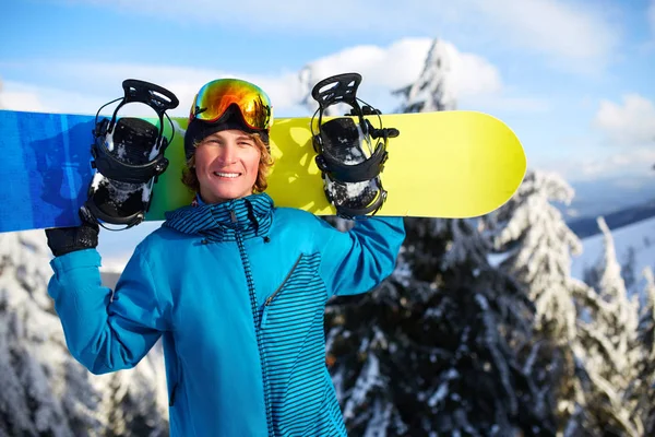 Sonriente snowboarder posando llevando snowboard sobre hombros en estación de esquí cerca del bosque antes de freeride backcountry y usando gafas reflectantes, colorido atuendo de moda. Equipo de snowboard moderno . —  Fotos de Stock