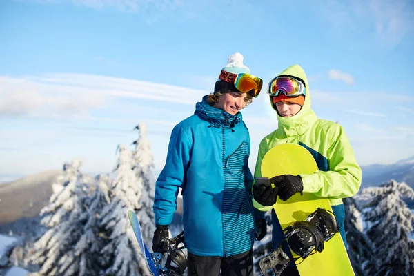 Dos snowboarders hablando y sosteniendo smartphones. Amigos mirando el teléfono celular y comunicándose en la red social compartiendo sus fotos en la aplicación con conexión a Internet con ropa de moda colorida . —  Fotos de Stock
