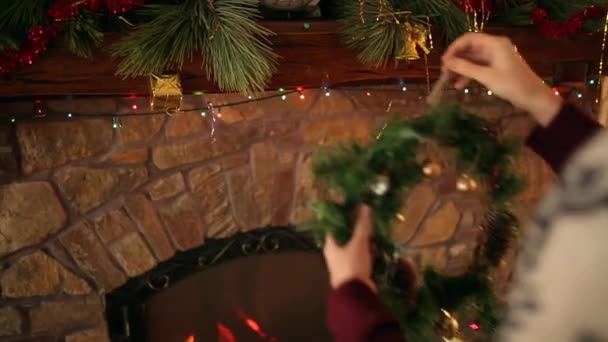 Человек в ножевом свитере висит рождественский венок над каменным подлинным камином, украшенным красочными гирляндскими огнями. Новогодние праздники подготовка и оформление. Смотреть в оба . — стоковое видео