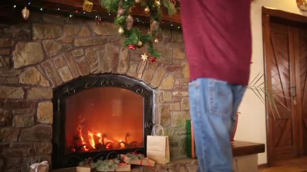 Man in kniited sweater hanging Christmas wreath above stone authentic fireplace decorated with colorful flashing garland lights. New Year holidays preparation and decoration. Medium view dolly shot. — Stock Video