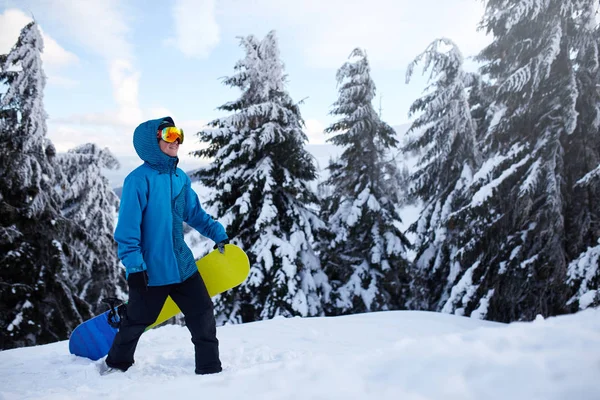 Snowboarder escalando a la cima de la montaña llevando su snowboard a través del bosque para freeride backcountry y usando gafas reflectantes, colorido traje de moda y pasamontañas en la estación de esquí. Deportes de invierno . —  Fotos de Stock