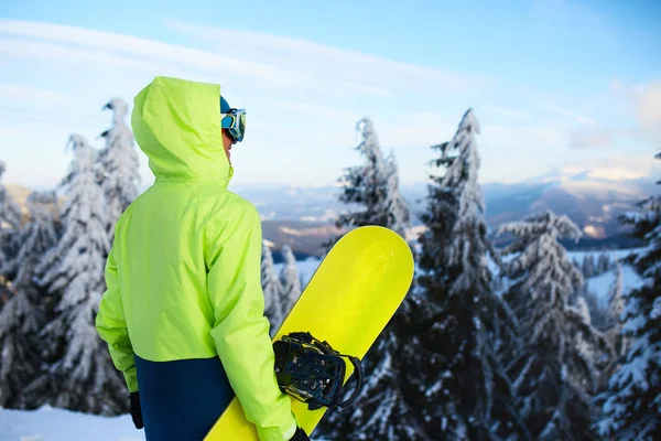 Vista posterior de snowboarder de pie con su tabla en el monte antes de la sesión freeride backcountry en el bosque. Hombre con snowboard mirando la estación de esquí. Jinete traje de moda de cal. —  Fotos de Stock