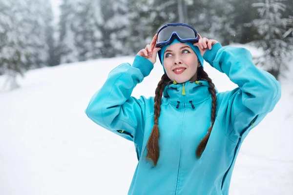 Mujer nórdica sonriente con coletas se pone gafas protectoras de esquí. Niña snowboarder tocando máscara en estación de esquí en nevadas cerca del bosque. Deportista elegante de ojos azules en traje de moda colorido . —  Fotos de Stock