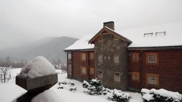 Houten vakantiehuis in bergdorp tijdens zware sneeuwval met naaldhout bos op de achtergrond. Sneeuw vallen op logboek chalets in skiresort. Koude frosty winterdag in de bergen. — Stockvideo