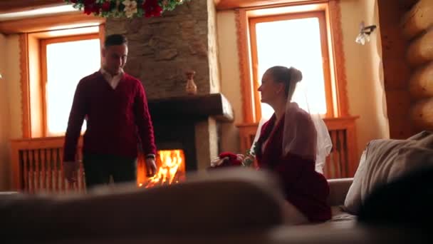 恋爱中的一对夫妇穿着红色毛衣在壁炉旁热身。新娘和新郎放松和亲吻温暖的火和温暖在原木木制小屋。冬季圣诞假期和蜜月概念. — 图库视频影像
