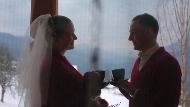 Bräutigam und Braut trinken Kaffee oder Tee aus Tassen auf dem Balkon eines Holzblockhauses im Dorf und küssen sich mit schneebedeckten Bergen und Hügeln im Hintergrund. Winter Hochzeit Flitterwochen Inspiration. — Stockvideo
