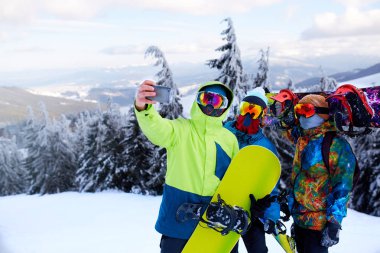 Üç snowboard kayak merkezi kameraya smartphone ile selfie alarak. Arkadaşlar sosyal ağ snowboards yakınındaki yansıtıcı gözlük, renkli moda ceket giyen orman ile paylaşım için fotoğraf çekimi