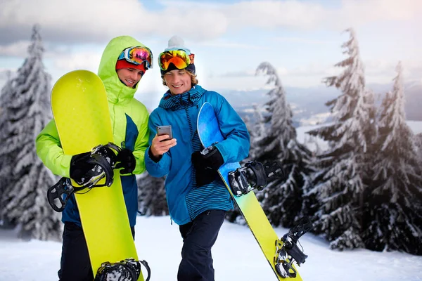 Dos snowboarders hablando y sosteniendo smartphones. Amigos mirando el teléfono celular y comunicándose en la red social compartiendo sus fotos en la aplicación con conexión a Internet con ropa de moda colorida . —  Fotos de Stock