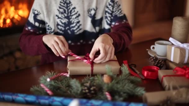 El hombre barbudo que se sienta y que ata el lazo a los regalos para Nuevo Año cerca de la chimenea. Tipo con sombrero de Navidad envolviendo cajas de regalo en papel añadiendo ramas de abeto, conos, caramelos de caña. Secuencia del paquete clip . — Vídeos de Stock