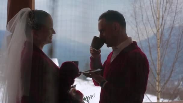 Groom et mariée boivent du café ou du thé dans des tasses sur un balcon en bois chalet en bois dans le village et baiser avec des montagnes couvertes de neige et des collines sur le fond. Mariage d'hiver lune de miel inspiration . — Video