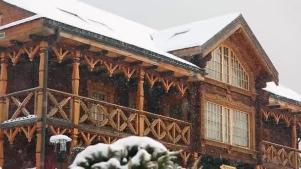 Close view of cottage with mezzanine in mountain village during heavy snowfall with russian style carved wooden facade. Snow falling on log chalets at ski resort. Cold frosty winter day. Slow motion. — Stock Video