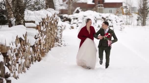 Casal de casamento jovem correndo, se divertindo de mãos dadas na aldeia resort de esqui com casas de madeira perto de logs. Inspiração de casamento de inverno. Noiva e noivo na lua-de-mel. Recém-casados de férias . — Vídeo de Stock