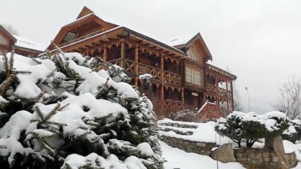 Wooden cottages in mountain village during heavy snowfall with coniferous forest on background. Snow falling on log chalets at ski resort with evergreen landscape design. Cold frosty winter day. — Stock Video