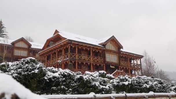 Cabañas de madera en pueblo de montaña durante las fuertes nevadas con bosque de coníferas en el fondo. Nieve cayendo en chalets de madera en la estación de esquí con un diseño de paisaje siempre verde. Frío día de invierno helado . — Vídeos de Stock