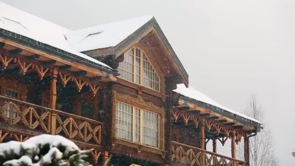 Close view of cottage with mezzanine in mountain village during heavy snowfall with russian style carved wooden facade. Snow falling on log chalets at ski resort. Cold frosty winter day. Slow motion. — Stock Video