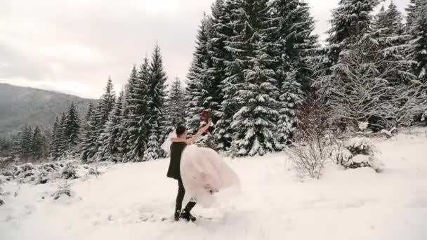 Groom spinning happy bride holding and spinning her in his hands in snow weather fir tree spruce forest during snowfall. Happy newlyweds have fun. Woman with bouquet. Valentines Day Christmas concept. — Stock Video