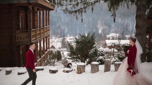 Brudgummen kommer till bruden, kramar och kyssa henne i snöig bergsby på ski resort. Romantiskt bröllop par i stickade marsala tröjor i låg under vinter snöfall. Jul och alla hjärtans dag. — Stockvideo