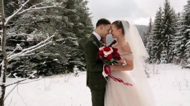 Yeni evliler damat ve gelin öpücük sarılmak ve birbirlerine ağır çekimde kar yağışı sırasında karlı herdem yeşil orman sıcak. . Kış ormanda genç bir çift. Kadın holding buket. Sevgililer günü ve Noel.
