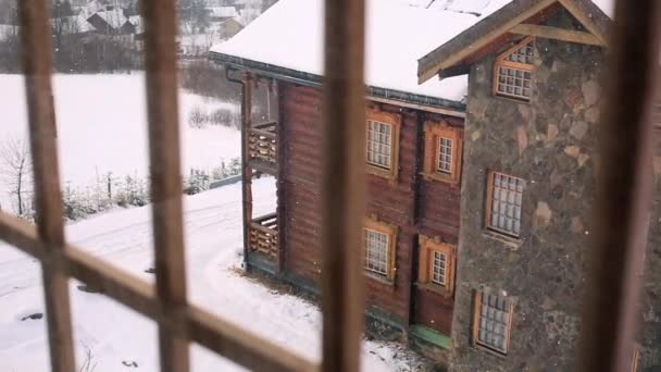 Via het venster weergave van houten huisje in bergdorp tijdens zware sneeuwval. Sneeuw vallen op logboek chalets met stenen schoorsteen muur in skiresort. Koude frosty winterdag in de bergen. — Stockvideo