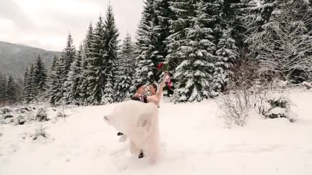 Bräutigam spinnt glückliche Braut hält und dreht sie in seinen Händen in Schnee Wetter Tannenwald bei Schneefall. glückliche Frischvermählte haben Spaß. Frau mit Strauß. Valentinstag-Weihnachtskonzept. — Stockvideo