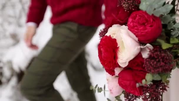 Bride carrying bouquet of flowers made of white and red roses. Young wedding couple walking in snowy forest during snowfall. Winter wedding inspiration. Low angle shot. Groom wearing marsala sweater. — Stock Video