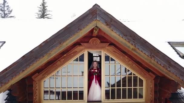 Drone camera flies away from beautiful bride standing on mezzanine of wooden chalet cottage revealing the mountain valley covered with spruce forest and snow. Fairy tale aerial from princess in castle — Stock Video