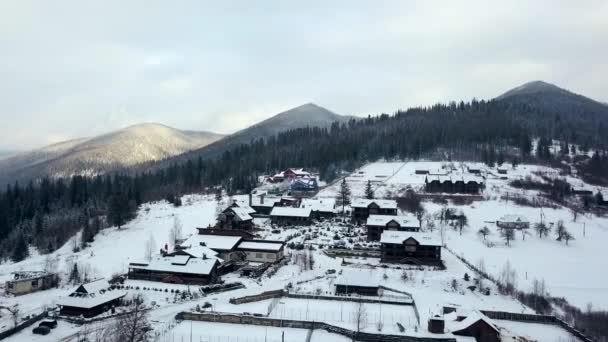 Αεροφωτογραφία του κατοικημένη περιοχή στο βουνό στο χειμώνα. Ορεινό χωριό κτίρια και σπίτια στις πλαγιές του λόφου χιονισμένη καλυμμένα με χιόνι. Στην εξοχή, στο δέντρο και πεύκα δάσος ελάτης. Εξοχικές κατοικίες του χιονοδρομικού. — Αρχείο Βίντεο