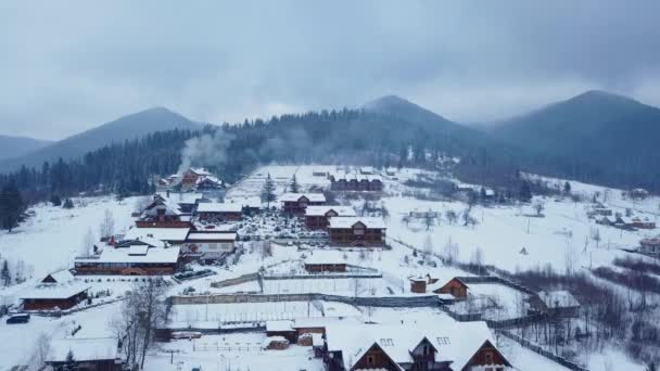겨울에 산에 살고 있는 지역의 항공 사진 산 마 건물 들과 눈 덮인 언덕 사면에 눈으로 덮여 있다. 시골, 전나무 나무와 소나무 숲입니다. 굴뚝에서 연기. — 비디오