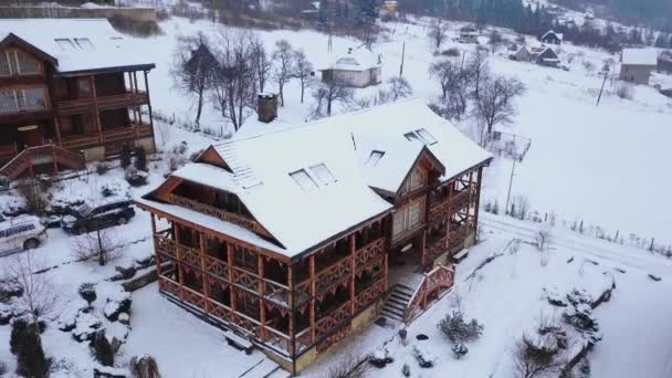 Aerial of wooden cottages in mountain village surrounded with coniferous forest. Drone view of chalets covered with snow at ski resort. Cold frosty winter day and snowfall in the mountains. — Stock Video