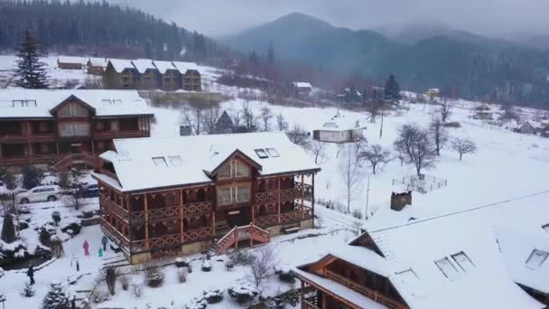 Ar de casas de madeira na aldeia montesa cercada de floresta conífera. Vista drone de chalés cobertos de neve na estância de esqui. Dia frio de inverno gelado e nevasca nas montanhas . — Vídeo de Stock