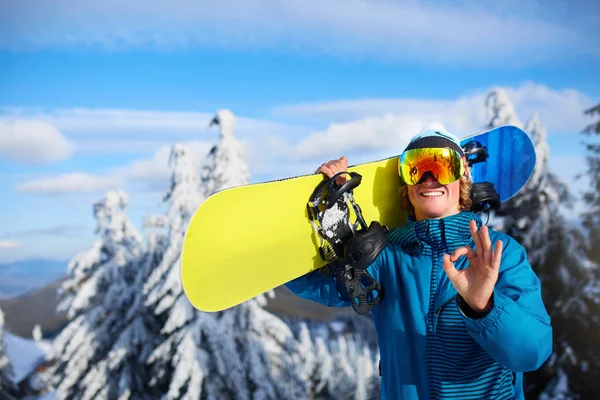 Sonriente snowboarder posando llevando snowboard sobre hombros en estación de esquí cerca del bosque antes de la sesión de freeride. Jinete mostrando signo OK usando gafas polarizadas. Equipo de snowboard moderno . —  Fotos de Stock