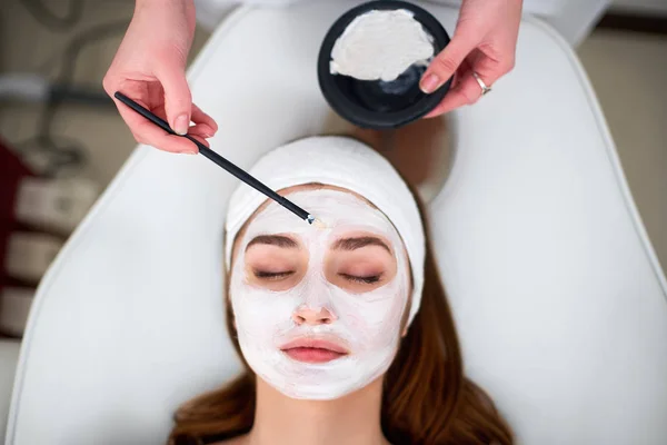 Hands of cosmetology specialist applying facial mask using brush, making skin hydrated and healthy. Attractive woman relaxing with closed eyes and enjoying skincare spa procedures. Beautician at work.