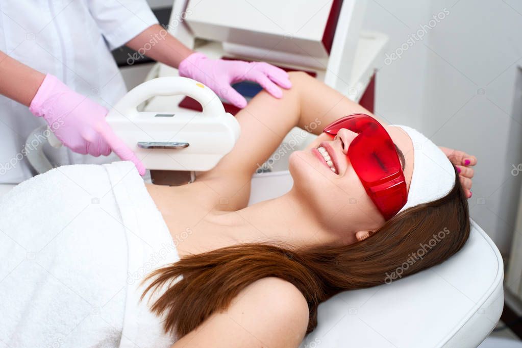 Beautician doctor doing elos hair removal depilation on attractive young woman armpits in beuty salon. Hardware aesthetic cosmetology. Laser epilation on underarms in clinic. Girl lying on couch.