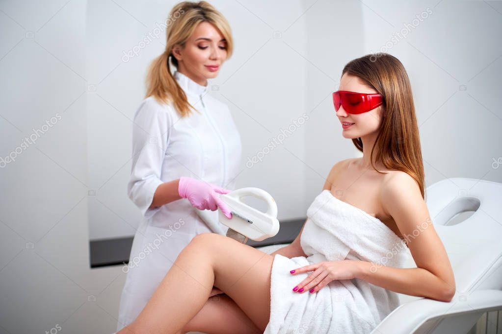 Beautician doctor doing elos hair removal depilation on attractive young woman legs in beuty salon. Hardware aesthetic cosmetology. Laser epilation on feet in clinic. Girl lying on couch.