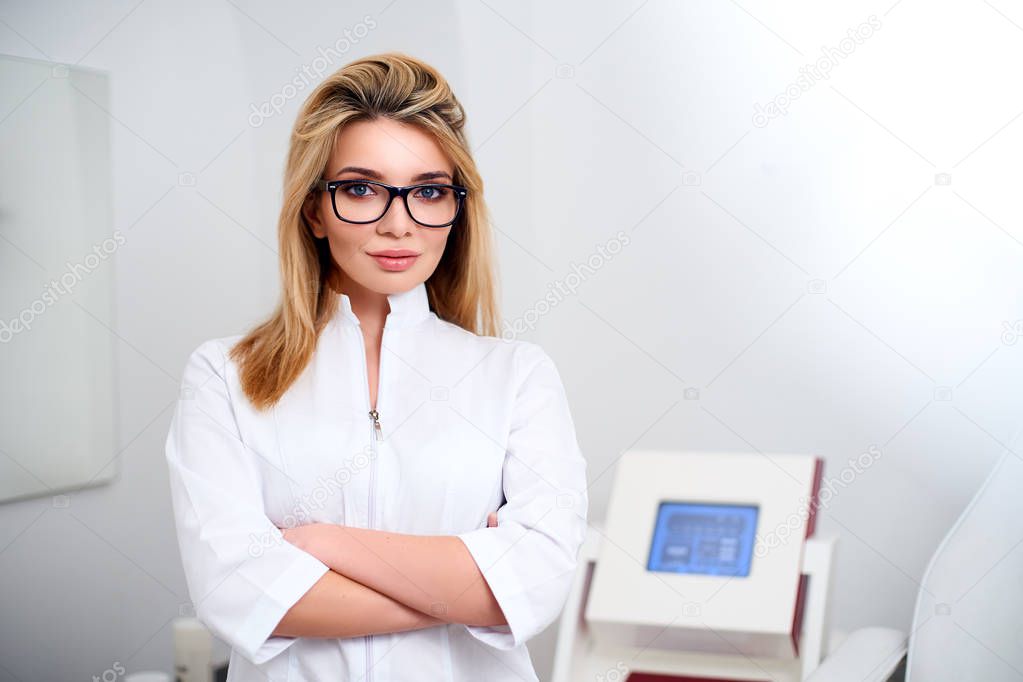Smiling confident female doctor with lab coat on standing in her office with medical hardware and patient chair on background. Practician woman wearing white hospital uniform and stylish glasses.