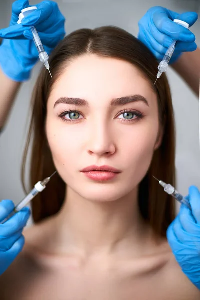 Manos de esteticistas sosteniendo jeringas alrededor de una cara de mujer impecable lista para la inyección en la clínica de cosmetología. Modelo femenino rodeado de jeringas de relleno. Concepto estándar de belleza moderna. Mesoterapia . — Foto de Stock