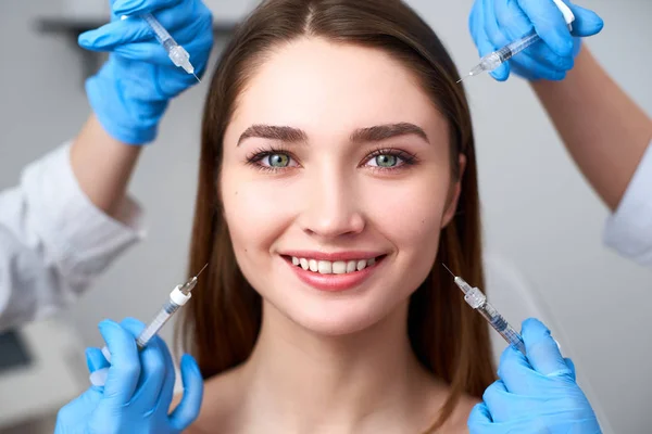 Handen van schoonheidsspecialisten houden spuiten rond vlekkeloze vrouw gezicht klaar voor injectie in cosmetologie kliniek. Vrouwelijk model omgeven met vulspuiten. Modern Beauty standaard concept. Mesotherapie. — Stockfoto