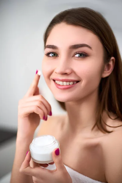 Smiling young mixed race woman applying face cream on her perfect skin. Model with flawless skin holding white jar of cream on blurred grey background in cosmetology clinic. — Stock Photo, Image
