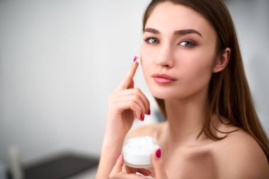 Onun mükemmel cilt üzerinde yüz kremi uygulayan gülümseyen genç karışık yarış kadın. Kozmetoloji kliniğinde bulanık gri arka plan üzerinde krem beyaz kavanoz tutan kusursuz cilt ile model.