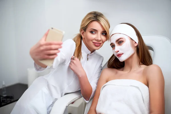 Sağlıklı ciltler için yüz maskesi uyguladıktan sonra müşteri ile selfie yapan güzellik blogger. Çekici kadınlar akıllı telefon ile fotoğraf yapma ve güzellik salonunda cilt bakımı spa prosedürleri zevk rahatlatıcı.