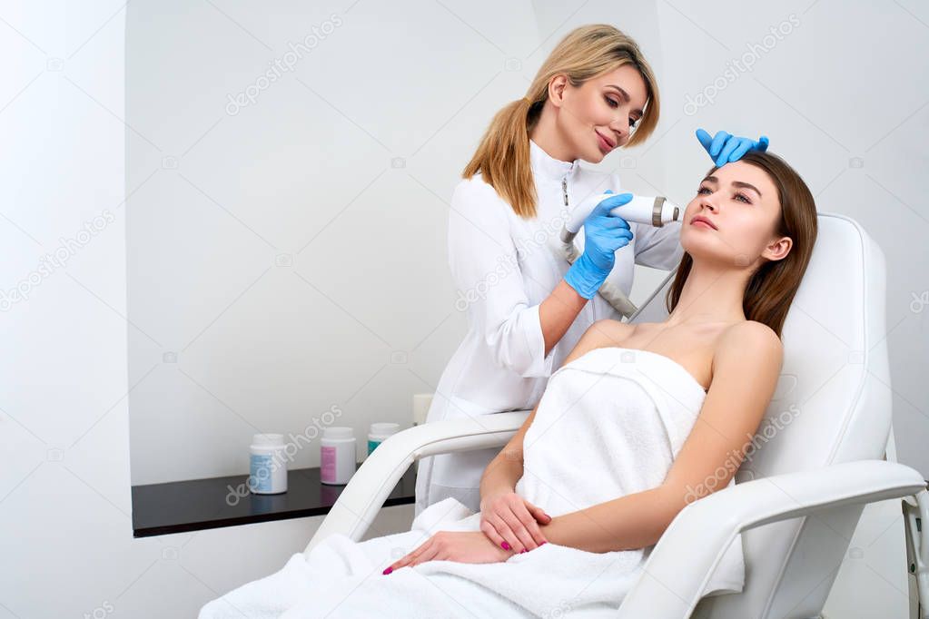 Pretty blonde beautician doctor doing rf-lifting procedure for woman laying in a beauty salon. Hardware cosmetology. Patient receiving electric facial massage. Skin rejuvenation and wrinkle smoothing.