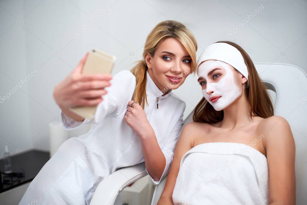 Beautician blogger making selfie with client after applying facial mask for healthy skin. Attractive women making photos with smartphone and relaxing enjoying skincare spa procedures in beauty salon.