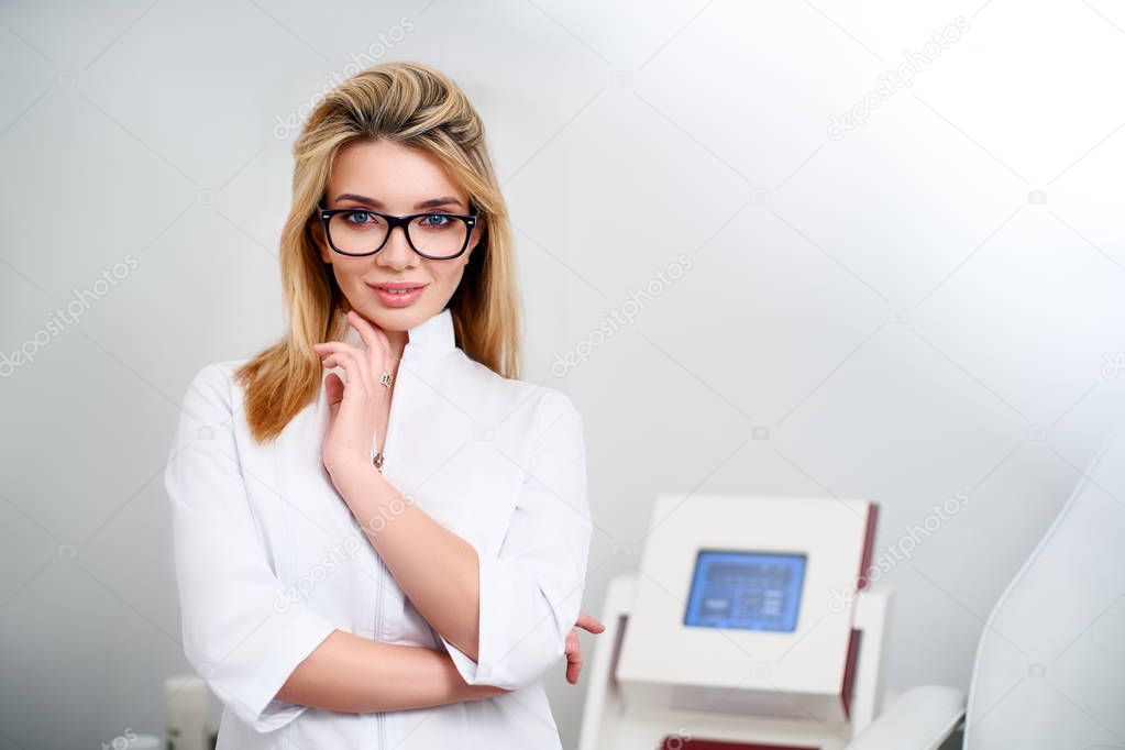 Smiling confident female doctor with lab coat on standing in her office with medical hardware and patient chair on background. Practician woman wearing white hospital uniform and stylish glasses.