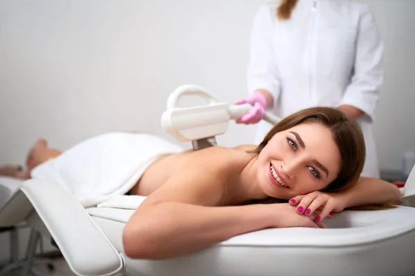 Young pretty smiling woman back laser epilation in beauty salon. Cosmetologist doing depilation treatment with elos device. Beautician hands in gloves holding hair removal cosmetology ipl hardware — Stock Photo, Image