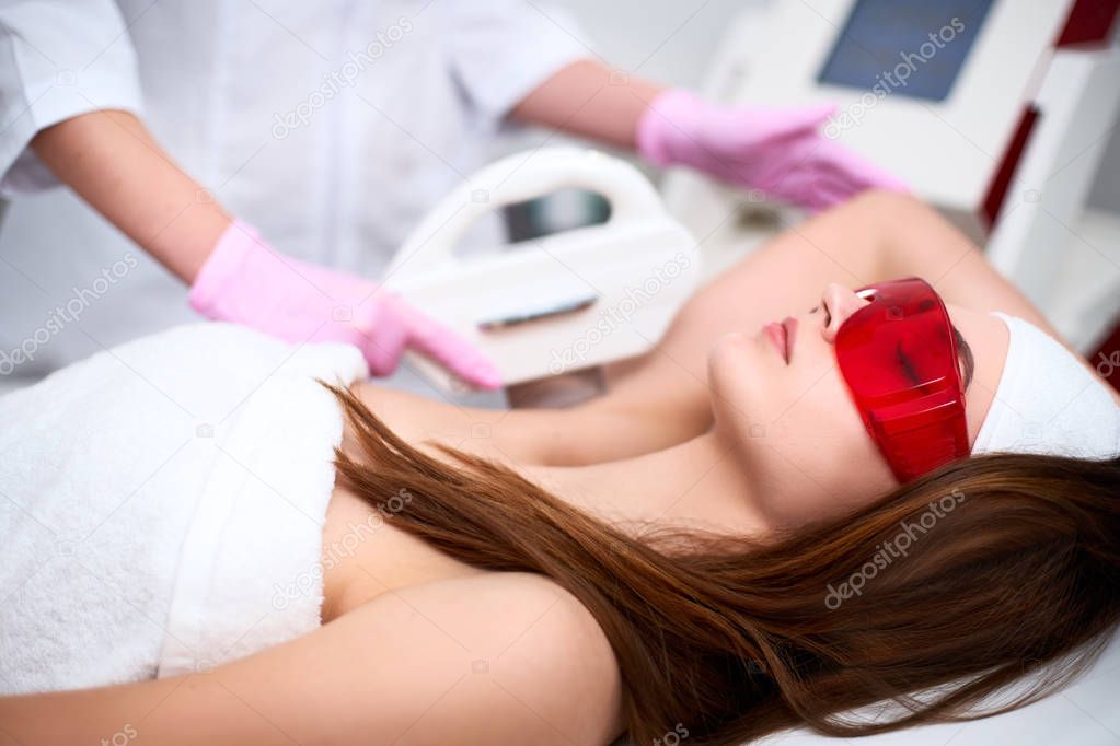 Beautician doctor doing elos hair removal depilation on pretty young woman armpits in beuty salon. Hardware aesthetic cosmetology. Laser epilation on underarms in clinic. Girl lying on couch.