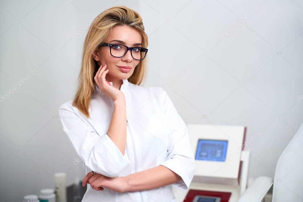 Smiling confident female beautician doctor in lab coat standing in her office with medical hardware and patient chair on background. Woman showing her flawless skin in hospital uniform and glasses.