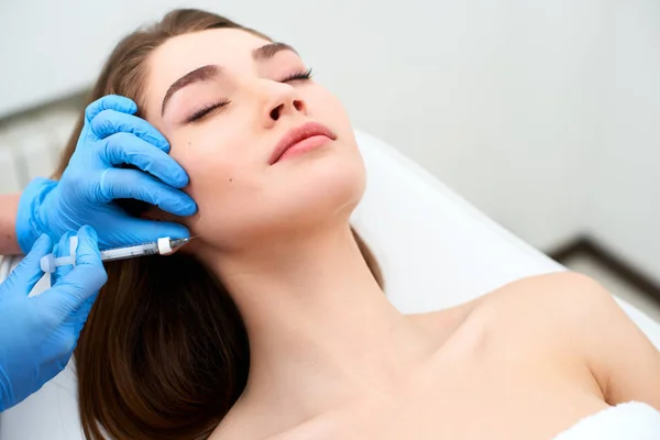 Médico esteticista con jeringa de relleno que hace la inyección a las papadas. Reducción de líneas maseteras y terapia de contorno facial. Tratamiento antienvejecimiento y lifting facial en clínica de cosmetología. Paciente acostado en silla —  Fotos de Stock