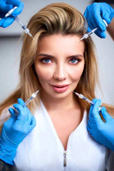 Hands of beauticians holding syringes around flawless woman face ready for injection in cosmetology clinic. Female model surrounded with filler syringes. Modern beauty standard concept. Mesotherapy.