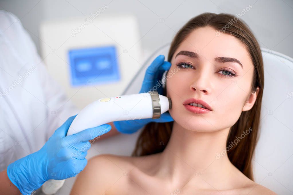 Beautician doctor doing rf-lifting procedure for flawless woman face laying in a beauty salon. Hardware cosmetology. Patient receiving electric facial massage. Skin rejuvenation and wrinkle smoothing.