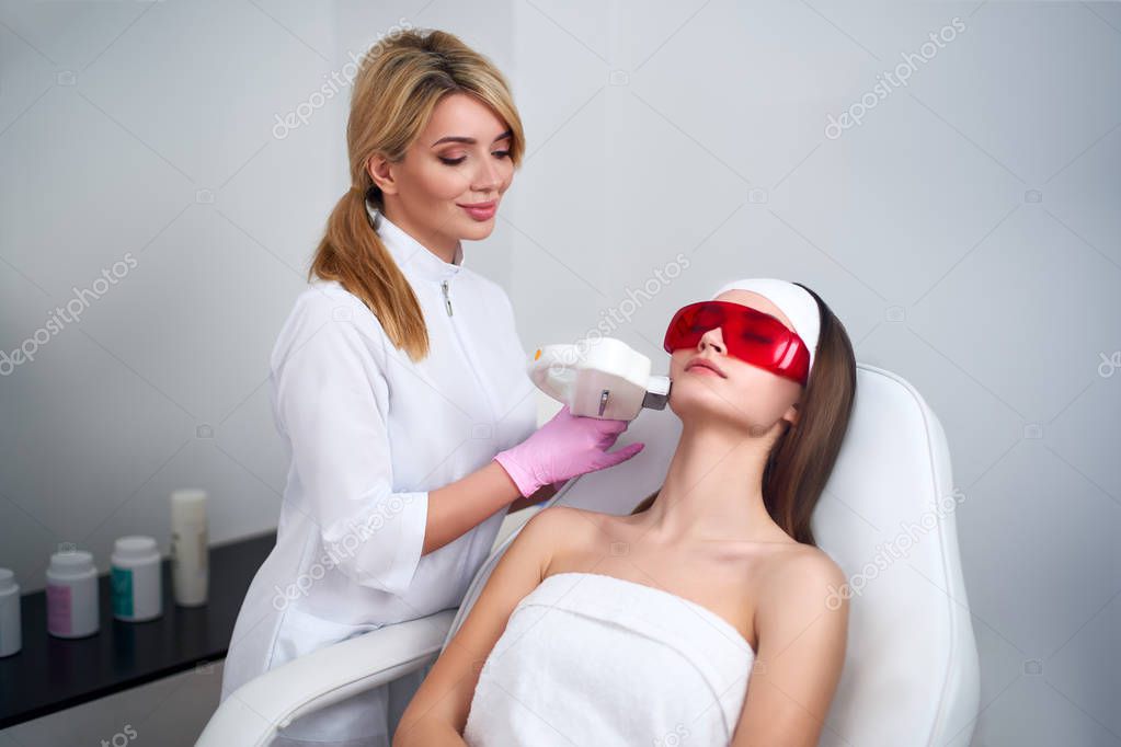 Beautician doctor doing laser RF rejuvenation for pretty young woman face at beauty salon. Elos epilation hair removal procedure. Aesthetic facial acne treatment skin care. Hardware ipl cosmetology